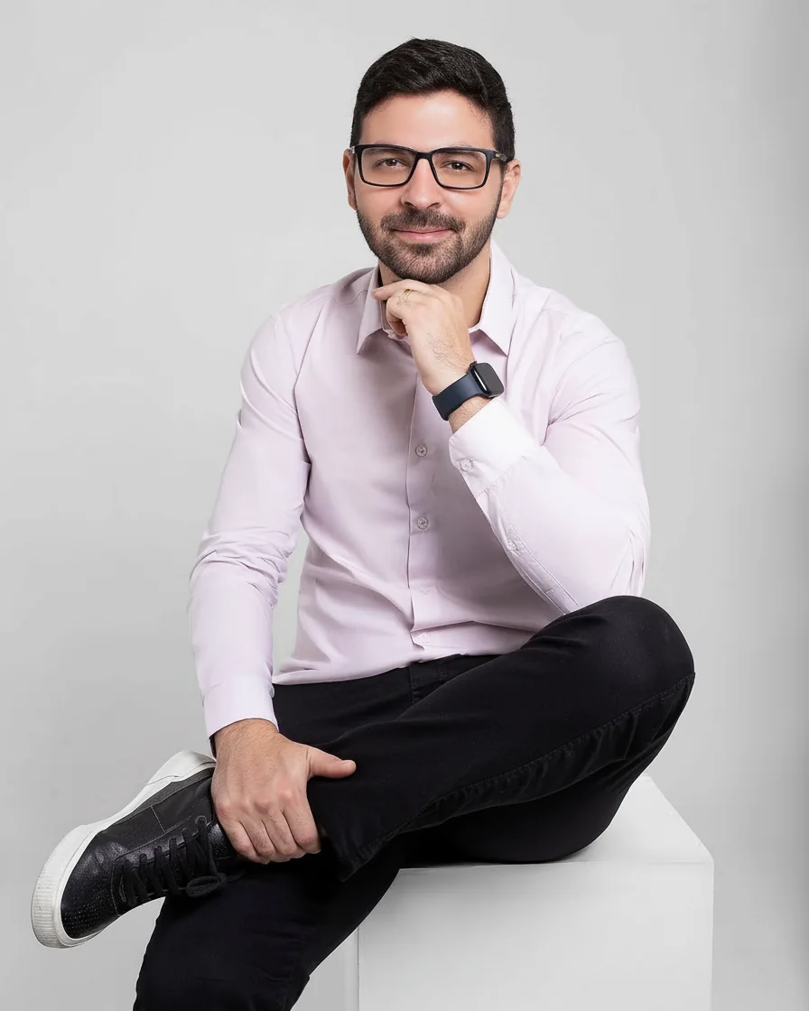 Dr. Henrique Moura sorrindo, sentado, de camisa clara e calça escura, sobre fundo cinza claro